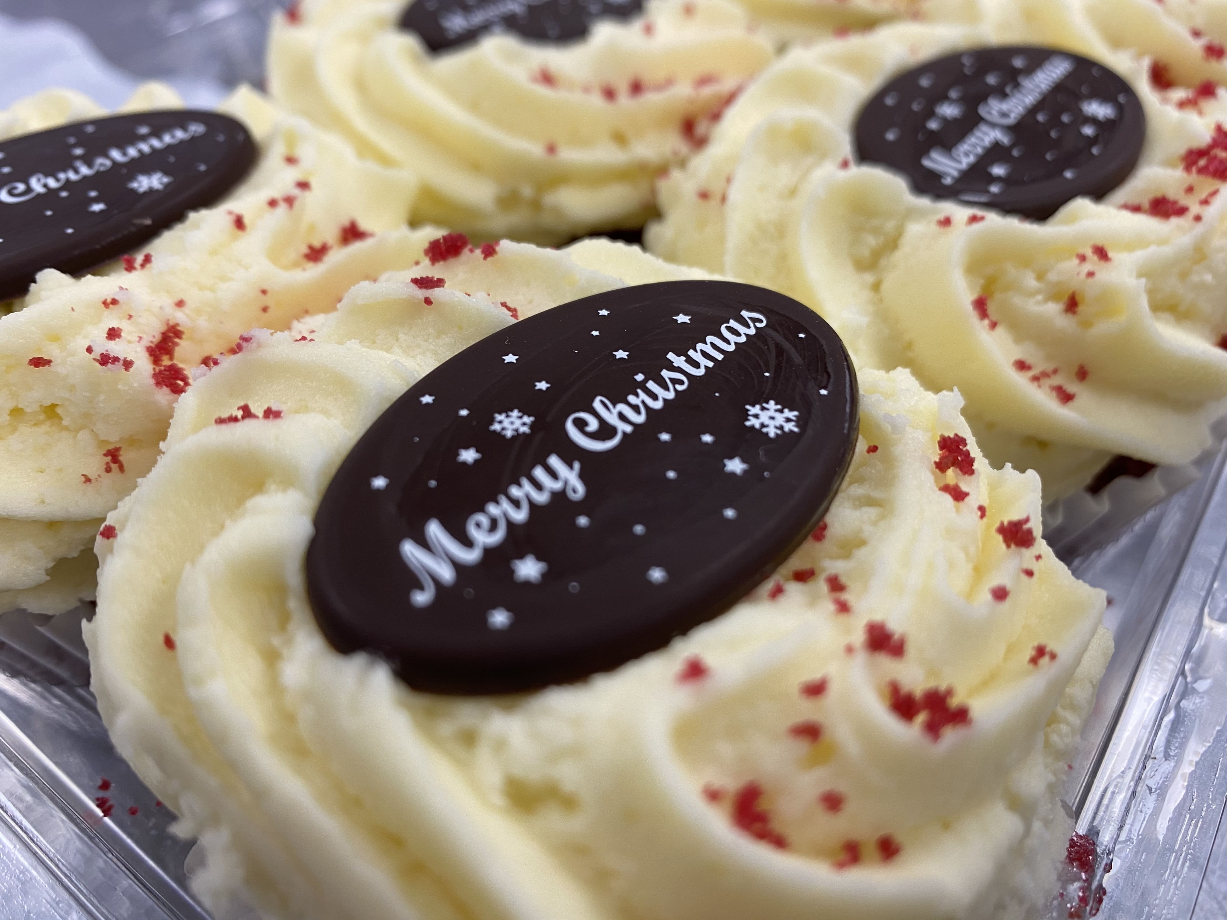 Close-up of Christmas-themed cookies with white swirled frosting and red sprinkles. Each cookie has a dark chocolate disc on top with 'Merry Christmas' written in white lettering along with small snowflake decorations, offering a delightful holiday dessert experience.